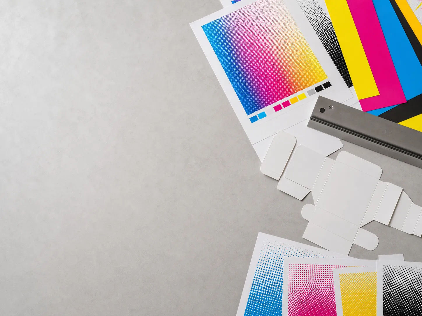CMYK කොළ, හිස් packaging හැඩතල, halftone සාම්පල් සහ යකඩ රූලක් තියෙන print-studio එකක උඩින් ගත්ත ආදර්ශන පින්තූරයක්.